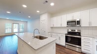 This contemporary single-floor residence offers a thoughtfully designed open-concept layout that seamlessly connects the living and kitchen areas. Rich hardwood-style flooring flows throughout the main living spaces, creating a warm and cohesive aesthetic.

The kitchen is a standout feature, showcasing white shaker cabinetry, granite countertops, and a classic subway tile backsplash. A central island with a built-in sink provides additional prep space and casual seating. A full suite of stainless steel appliances — including a refrigerator, electric range, built-in microwave, and dishwasher — completes the space with a clean, modern finish.

The living area is bright and open, enhanced by recessed lighting and a glass-panel door that invites natural light while offering direct outdoor access. Bedrooms feature plush carpeting, ceiling fans, and large windows with exterior views. Generous closet space with built-in shelving ensures ample storage.

The bathroom is finished with floor-to-ceiling white tile surrounding a soaking tub, delivering a clean and timeless look. Throughout the residence, crisp white walls, recessed lighting, and consistent finishes create a bright, move-in-ready environment that balances comfort with modern style.