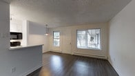 This single-floor residence offers a practical and well-appointed layout designed for comfortable everyday living. The open living area serves as the heart of the space, showcasing rich, dark hardwood flooring that runs throughout and provides a warm contrast to the bright white walls and textured ceiling. Natural light flows generously into the room through multiple windows fitted with horizontal blinds, illuminating the space and creating an inviting atmosphere. A white paneled front entry door provides a welcoming focal point, flanked by windows that offer views of the surrounding greenery outside. Baseboard heating units run along the exterior walls, ensuring efficient warmth throughout the cooler months. A partial kitchen counter or peninsula is visible at the edge of the living area, hinting at an open-concept connection between the living and kitchen spaces. A pendant light fixture hangs above, adding a subtle decorative touch to the overall clean and neutral aesthetic. The textured popcorn ceiling adds a classic residential character to the space. Overall, the unit presents a tidy, move-in-ready environment with functional finishes and a bright, airy feel suited to a range of living arrangements.