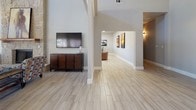 This single-floor space is defined by its dramatic architectural design and open layout. The main living area commands attention with soaring vaulted ceilings, exposed structural beams, and light wood-tone tile flooring that flows seamlessly throughout. A floor-to-ceiling natural stone fireplace serves as the centerpiece of the living room, paired with warm artwork and eclectic, comfortable seating that adds character to the space.

The dining area is bathed in natural light, thanks to a striking arched window and French doors that open to an outdoor area, creating a bright and airy atmosphere. A rustic wooden dining table and chairs add warmth and a sense of casual elegance to the room.

The kitchen features gray cabinetry, crisp white countertops, a patterned geometric mosaic tile backsplash, and black appliances. A window above the sink provides additional natural light, keeping the space feeling open and functional.

A well-lit corridor connects the various areas, lined with wall sconces and accented with artwork. A cozy seating nook off the hallway, furnished with studded leather chairs and a woven rug, offers an intimate retreat. Throughout the space, neutral wall tones, consistent flooring, and carefully chosen décor create a cohesive, inviting ambiance suited for both relaxing and entertaining.
