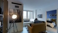 This single-floor space showcases a contemporary urban aesthetic, blending clean architectural lines with thoughtfully curated design elements. The living area is anchored by large windows that frame impressive city skyline views, flooding the interior with natural light and creating an open, airy atmosphere. A built-in window seat beneath the windows offers a comfortable vantage point to take in the urban scenery, accented with neatly arranged throw pillows in muted tones. The light hardwood flooring and white walls serve as a neutral backdrop that allows the décor to take center stage. A bold black-and-white photographic mural of a suspension bridge commands attention on one wall, contributing a dramatic, artistic focal point to the space. Complementing this, a framed monochrome portrait photograph adds further visual interest. A dark, multi-drawer storage cabinet grounds the room and provides functional storage, while a warm mustard-toned sofa introduces a pop of color. An industrial-style spotlight on a tripod stand reinforces the space's modern, editorial character. Together, these elements create a sophisticated and visually dynamic environment.