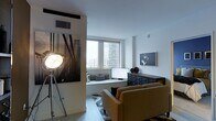 This single-floor space showcases a contemporary urban aesthetic, blending clean architectural lines with thoughtfully curated design elements. The living area is anchored by large windows that frame impressive city skyline views, flooding the interior with natural light and creating an open, airy atmosphere. A built-in window seat beneath the windows offers a comfortable vantage point to take in the urban scenery, accented with neatly arranged throw pillows in muted tones. The light hardwood flooring and white walls serve as a neutral backdrop that allows the décor to take center stage. A bold black-and-white photographic mural of a suspension bridge commands attention on one wall, contributing a dramatic, artistic focal point to the space. Complementing this, a framed monochrome portrait photograph adds further visual interest. A dark, multi-drawer storage cabinet grounds the room and provides functional storage, while a warm mustard-toned sofa introduces a pop of color. An industrial-style spotlight on a tripod stand reinforces the space's modern, editorial character. Together, these elements create a sophisticated and visually dynamic environment.