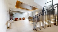 Located on Santa Monica Boulevard in Los Angeles, this refined commercial lobby presents a polished first impression through its thoughtful architectural design and high-end finishes. The spacious hallway is anchored by large-format cream-toned floor tiles that run seamlessly throughout, lending a sense of openness and continuity to the space.

A striking wood-paneled tray ceiling with recessed and track lighting serves as a central design feature, adding warmth and depth to the otherwise crisp, white interior. The reception desk, clad in brushed bronze-toned metal with a dark countertop, provides a sleek and functional focal point. Adjacent to the reception area, a comfortable seating arrangement with curved, earth-toned lounge chairs invites visitors to relax.

A glass-partitioned section allows natural light to flow inward from street-facing windows, enhancing the bright and airy ambiance. A sculptural staircase with dark metal railings leads to upper levels, while a stainless steel elevator offers convenient vertical access. Decorative accents — including a stone console table, artful branch arrangements, and a richly veined green marble wall panel — add curated elegance throughout. The overall atmosphere is modern, professional, and welcoming.