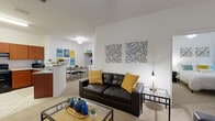 Located at 3205 NW 83rd St in Gainesville, FL, this single-floor unit offers a well-designed, open-concept layout that seamlessly connects the living, dining, and kitchen areas. The spacious living room features plush carpeting, a ceiling fan, and large windows that invite natural light and provide views of the surrounding landscape. A wall-mounted flat-screen TV and ample seating make it an ideal space for relaxing or entertaining.

The kitchen is thoughtfully appointed with warm wood cabinetry, granite-style countertops, a full suite of black appliances including a range, refrigerator, and dishwasher, and tile flooring. A peninsula counter opens toward the dining area, creating an easy flow for everyday living. A dedicated dining space sits just off the kitchen, providing a practical area for meals.

The bedrooms are carpeted and well-lit, each featuring ceiling fans, generous closet space, and windows with draping curtains that offer privacy while allowing natural light. The rooms comfortably accommodate a full bedroom setup, including space for a desk — ideal for studying or working from home.

The bathroom is clean and functional, featuring tile flooring, a granite-topped vanity with wood cabinetry, and a tub-shower combination. Overall, the unit presents a bright, comfortable, and practical living environment.