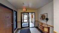 This single-floor space presents a functional and modestly decorated lobby area designed to serve residents or occupants efficiently. The entrance corridor features tile flooring accented by welcome mats, with dark-framed glass doors providing access to the outdoors. Natural light filters in through the glazed entry doors, offering views of the surrounding exterior. A textured ceiling with standard overhead lighting and an illuminated exit sign contribute to the utilitarian character of the space.

Along one wall, a bank of individual metal mailboxes provides secure mail storage, positioned beneath a large, decorative open-face clock with Roman numerals and a Paris-themed design, adding a subtle stylistic touch to an otherwise practical area. A wooden credenza with ornate detailing sits nearby, complemented by a seasonal wreath displayed on the wall, lending a welcoming feel to the entryway.

Wood chair rail molding runs along the walls, providing visual definition between the lower and upper portions of the corridor. Sconce lighting adds warm illumination to the mailbox area, where a chair with a patterned cushion offers a modest seating option. Overall, the space balances practicality with light decorative touches, creating a functional and approachable common entry area.