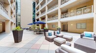 Located at 755 E Broad St in Athens, Georgia, this expansive outdoor courtyard offers a generously sized living area designed for both relaxation and social gathering. The space is framed by a multi-story building facade featuring ornate decorative railings adorned with repeating medallion motifs on each level, giving the courtyard a distinctly refined architectural character. Large structural columns and warm-toned stucco walls define the perimeter, while wall-mounted lantern-style sconces provide ambient lighting throughout.

The courtyard is appointed with contemporary outdoor furnishings, including modular wicker lounge chairs and sofas with plush white cushions accented by blue and teal throw pillows. A dedicated dining zone features a table and chairs shaded by a bold navy market umbrella, ideal for al fresco meals. Tall, angular planters in slate gray are strategically placed throughout the space, housing lush ornamental trees and flowering plants that introduce natural texture and greenery.

The tiled floor features a pattern of neutral and earth-toned pavers that define different zones within the courtyard. A glass curtain wall on one side of the building tower adds a modern architectural contrast. The overall ambiance is polished, open-air, and welcoming, offering a well-curated communal outdoor retreat.
