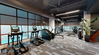 Located at 4510 Beech St in Norwood, Ohio, this contemporary fitness center spans 1,640 square feet on a single floor, offering a comprehensive and well-appointed workout facility. The space is defined by its industrial-modern aesthetic, featuring exposed dark-painted structural ceilings with visible beams and ductwork, complemented by sleek linear LED lighting that illuminates the entire floor plan.

Floor-to-ceiling grid-pane windows line the perimeter, flooding the cardio zone with natural light and providing open views of the surrounding Norwood cityscape. This area is equipped with multiple treadmills, ellipticals, a stair climber, and a rowing machine, all featuring integrated display screens. Large ceiling fans enhance air circulation throughout.

The strength training zone includes a full rack of dumbbells displayed on tiered racks, a multi-functional cable and Smith machine system, and various weight benches and cable attachment equipment. A large wall mirror expands the sense of space and supports proper form during workouts.

A dedicated utility area provides a wall-mounted TV screen, a water station with a built-in dispenser, and additional specialty equipment. Patterned, durable carpet tiles cover the floor throughout, adding texture and comfort underfoot. The overall ambiance is sleek, modern, and energizing, designed to support a full range of fitness activities.