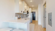 This single-floor unit showcases a clean, contemporary design throughout, with light hardwood-style flooring and crisp white walls creating a bright, cohesive aesthetic. The kitchen is a standout feature, finished with white shaker-style cabinetry accented by matte black hardware and a soft blue-grey subway tile backsplash. Light stone countertops extend into a compact peninsula, providing additional prep and seating space. Stainless steel appliances, including a refrigerator, dishwasher, over-the-range microwave, and sink with a modern faucet, equip the kitchen fully. A hallway leads from the kitchen to the entry, finished with a dark front door that contrasts the overall light palette. The bedroom is comfortably scaled, featuring light-filled windows with sleek dark frames that frame views of the surrounding urban streetscape and a private balcony access. A wall-mounted HVAC unit ensures year-round climate control. The bathroom is finished in an all-white palette, with a quartz-topped vanity, a large mirror, and a full tub-shower combo with white tile surround. In-unit stacked laundry is also present, adding functional convenience to the space.