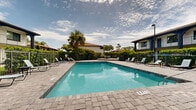 Located at 1250 Woodcrest Dr in Daytona Beach, Florida, this outdoor pool area offers a welcoming retreat designed for relaxation and recreation in a classic Florida setting. The centerpiece is a large, clearly defined swimming pool with crisp, blue water and stainless steel entry railings, set atop an expansive paver stone deck that provides ample space for lounging and socializing. Numerous sling-style lounge chairs and seating arrangements are positioned around the perimeter, allowing guests to enjoy the abundant Florida sunshine. A poolside utility structure with a terra cotta tile roof provides shade and architectural character, complementing the Mediterranean-inspired design of the surrounding multi-story buildings, which feature arched balconies, tile rooflines, and stucco exteriors. The entire pool area is enclosed by a black iron security fence, beyond which dense tropical landscaping — including mature palms, lush hedgerows, and a variety of flowering shrubs — creates a sense of privacy and seclusion. A raised seating platform at one end of the deck offers an elevated vantage point, while a shaded umbrella table provides an additional spot to unwind. The overall ambiance is that of a well-maintained, resort-style outdoor space that captures the relaxed, sun-drenched spirit of Daytona Beach.