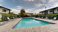 Located at 1250 Woodcrest Dr in Daytona Beach, Florida, this outdoor pool area offers a welcoming retreat designed for relaxation and recreation in a classic Florida setting. The centerpiece is a large, clearly defined swimming pool with crisp, blue water and stainless steel entry railings, set atop an expansive paver stone deck that provides ample space for lounging and socializing. Numerous sling-style lounge chairs and seating arrangements are positioned around the perimeter, allowing guests to enjoy the abundant Florida sunshine. A poolside utility structure with a terra cotta tile roof provides shade and architectural character, complementing the Mediterranean-inspired design of the surrounding multi-story buildings, which feature arched balconies, tile rooflines, and stucco exteriors. The entire pool area is enclosed by a black iron security fence, beyond which dense tropical landscaping — including mature palms, lush hedgerows, and a variety of flowering shrubs — creates a sense of privacy and seclusion. A raised seating platform at one end of the deck offers an elevated vantage point, while a shaded umbrella table provides an additional spot to unwind. The overall ambiance is that of a well-maintained, resort-style outdoor space that captures the relaxed, sun-drenched spirit of Daytona Beach.