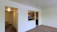 This single-floor space is designed with a practical and functional layout. A carpeted staircase with a dark wood handrail and crisp white risers provides access between levels, featuring neutral-toned, textured carpeting that adds a sense of warmth and comfort underfoot.

The main living area is finished with wall-to-wall carpeting in a muted tone, creating a cohesive and inviting atmosphere. A defining architectural feature is the pass-through window connecting the living area to the kitchen, allowing for an open, communicative flow between the two spaces. The kitchen is equipped with warm wood cabinetry that extends to the upper and lower portions of the walls, providing ample storage. A gas range and additional appliances are visible, along with a countertop that runs the length of the kitchen.

The surrounding walls are painted a clean, bright white, enhancing the sense of openness throughout. A hallway visible from the living area suggests additional rooms beyond, with sliding doors providing closet or storage access. Overall, the space presents a clean, straightforward design with practical finishes suited for everyday living.