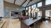 This expansive clubroom in Bryan, Texas is defined by its dramatic vaulted ceiling with exposed dark wood beams that rise to impressive heights, creating an open, airy atmosphere throughout the single-story space. Light wood-look tile flooring in a herringbone pattern flows seamlessly across the main areas, complementing the warm, neutral color palette.

A towering stone-clad fireplace serves as a striking architectural centerpiece, extending from floor to ceiling and anchoring one of several distinct seating zones. Multiple lounge areas are furnished with curved sectional sofas, upholstered accent chairs, and eclectic décor, offering flexible gathering options. A full-size billiards table provides a recreational focal point, positioned beneath stylish pendant lighting clusters with frosted globe shades.

A built-in wet bar and coffee station, finished with wood cabinetry and a tile backsplash, is paired with a wall-mounted flat-screen television, creating a comfortable media lounge area. A smaller dining nook with a round table and upholstered chairs offers intimate seating. Clerestory windows along the upper walls flood the interior with abundant natural light, while French doors provide views of the surrounding outdoor environment. A glass-walled conference or business room is also accessible from the main space.