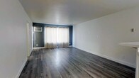 This single-floor space offers a clean, modern interior defined by thoughtful design choices and practical features. The main living area is spacious and open, showcasing wide-plank, grey-toned hardwood-style flooring that extends throughout, providing a sleek and contemporary foundation. The walls feature a two-tone design, with crisp white upper walls complemented by a bold dark grey accent wall that anchors the room and adds visual depth. Sheer curtains frame what appears to be a sliding glass door, allowing diffused natural light to filter into the space while offering access to an outdoor area. A wall-mounted air conditioning unit is conveniently positioned on the accent wall, ensuring climate control without occupying floor space. The white baseboards offer a clean contrast against the flooring, and the smooth, flat ceiling contributes to the overall minimalist aesthetic. The layout is open and unobstructed, providing flexible options for furniture arrangement and room configuration. Overall, the space presents a fresh, move-in-ready environment with a neutral palette that suits a variety of interior styles.