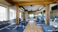 This single-floor fitness center combines rustic architectural charm with practical functionality. Exposed wooden beams and posts add a warm, lodge-inspired character to the space, contrasting nicely with the olive-toned walls and light hardwood-style flooring that runs throughout. Ceiling fans help maintain comfortable airflow during workouts.

The space is thoughtfully organized into distinct exercise zones. A free weights area along one side features a well-stocked dumbbell rack and a weight bench atop protective floor mats. On the opposite side, a row of cardio machines — including ellipticals and treadmills — is neatly arranged, benefiting from natural light streaming through multiple windows fitted with blinds. Large wall-mounted mirrors amplify the sense of space and allow users to monitor their form.

Overhead fluorescent lighting supplements the natural light, ensuring the space remains bright at all times. A wall-mounted television provides entertainment options during workouts. A solid wood interior door provides access, adding to the rustic aesthetic. The overall ambiance is inviting yet functional, balancing a cozy, natural design sensibility with a comprehensive fitness environment.