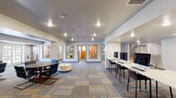 This well-appointed single-floor community space is thoughtfully designed to support both collaborative and focused work. The open main area is anchored by a patterned, multi-tone carpet that adds visual warmth and texture throughout. Recessed lighting illuminates the entire floor, creating a bright and welcoming ambiance.

The central lounge area features a round table surrounded by sleek, modern chairs with a cylindrical ottoman at its center, offering a comfortable setting for casual meetings or group gatherings. Along one wall, a built-in counter hosts a row of desktop computers on bar-height surfaces with tall chairs — a fully equipped workstation area ideal for productivity.

Framed artwork displayed on the walls adds a polished, curated touch to the space. A dedicated seating alcove with blue upholstered chairs and a round table provides a quieter setting for smaller discussions, complemented by a window that brings in natural light.

At the rear of the main area, a corridor leads to several private rooms, each distinguished by colorful accent walls — including a bold orange room and a green-tinted room — furnished with small tables and chairs suited for focused study or intimate meetings. The overall design balances open communal zones with accessible private spaces, making this a versatile and functional environment.