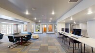 This well-appointed single-floor community space is thoughtfully designed to support both collaborative and focused work. The open main area is anchored by a patterned, multi-tone carpet that adds visual warmth and texture throughout. Recessed lighting illuminates the entire floor, creating a bright and welcoming ambiance.

The central lounge area features a round table surrounded by sleek, modern chairs with a cylindrical ottoman at its center, offering a comfortable setting for casual meetings or group gatherings. Along one wall, a built-in counter hosts a row of desktop computers on bar-height surfaces with tall chairs — a fully equipped workstation area ideal for productivity.

Framed artwork displayed on the walls adds a polished, curated touch to the space. A dedicated seating alcove with blue upholstered chairs and a round table provides a quieter setting for smaller discussions, complemented by a window that brings in natural light.

At the rear of the main area, a corridor leads to several private rooms, each distinguished by colorful accent walls — including a bold orange room and a green-tinted room — furnished with small tables and chairs suited for focused study or intimate meetings. The overall design balances open communal zones with accessible private spaces, making this a versatile and functional environment.