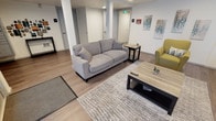 Located at 7911 5th Ave NE in Seattle, WA, this building lobby offers a clean, modern aesthetic designed to create a welcoming first impression. The space features light-toned hardwood-style flooring that extends throughout, contributing to an open and cohesive feel. A comfortable lounge area is centrally arranged, anchored by a plush gray sofa and complemented by a chartreuse accent chair, a wooden coffee table, and a side table — all set atop a neutral area rug that defines the seating zone. Contemporary artwork on the white walls adds a touch of color and visual interest to the otherwise understated palette. Recessed lighting provides consistent, even illumination throughout the space. Multiple interior doors provide access to additional areas of the building, including what appears to be utility and common-use rooms, all finished in a consistent white that maintains the space's clean, modern character. A wall-mounted control panel and signage indicate building systems are conveniently accessible within the lobby. Overall, the space balances functionality with a comfortable, inviting atmosphere suited for residents and visitors alike.