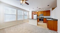 This single-floor residence is designed with an open-concept layout that seamlessly connects the living and kitchen areas. The living space features soft, neutral-toned carpeting and a ceiling fan with integrated lighting, creating a comfortable and functional environment. Multiple windows fitted with white blinds allow generous natural light to fill the room, contributing to an airy, welcoming atmosphere.

The kitchen is well-appointed with warm honey-toned cabinetry offering ample storage, complemented by striking dark granite countertops that extend across both the perimeter surfaces and a central peninsula. A complete suite of stainless steel appliances—including a refrigerator, electric range, built-in microwave, and dishwasher—provides a polished, modern aesthetic. Track lighting overhead ensures the workspace is thoroughly illuminated. Light ceramic tile flooring grounds the kitchen, creating a clean visual contrast to the adjacent carpeted living area.

A separate carpeted room, also equipped with a ceiling fan, serves as a versatile private space suitable for use as a bedroom or home office. The bathroom is cleanly finished with neutral tile flooring, a full soaking tub with a tiled shower surround in crisp white, and wood-toned cabinetry for additional storage. Overall, the space maintains a cohesive, neutral palette throughout.