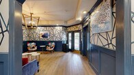 Located at 200 College Pl in Norfolk, Virginia, this thoughtfully designed lounge offers a compelling blend of classic architectural details and contemporary interior design. The space is unified by a striking geometric wallpaper pattern — bold navy lines set against a warm cream background — that wraps the walls and creates a dramatic, visually cohesive atmosphere throughout. Deep navy wainscoting lines the lower walls, complementing the decorative crown molding and dark-trimmed archways that add a sense of architectural refinement. Wide-plank hardwood flooring runs the length of the space, lending warmth and continuity underfoot. A seating area features a plush navy velvet sofa accented with red throw pillows, multicolored accent chairs, and marble-topped side tables, creating an inviting environment for conversation and relaxation. A navy-painted vintage display cabinet with brass hardware and a red interior serves as a standout decorative focal point. Recessed lighting and a brass chandelier provide layered illumination, while multiple French doors and windows allow natural light to filter in and offer views of the outdoor surroundings. A decorative archway elegantly connects the two areas of the lounge, enhancing the sense of flow. Original artwork is displayed throughout, adding personality and a curated gallery-like quality to the overall design.