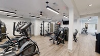 Located at 3400 Kimball Bridge Rd in Alpharetta, GA, this fitness center offers a thoughtfully designed exercise space spread across a single expansive floor. The room features light wood-look flooring, crisp white walls, and a clean, modern aesthetic that creates an energizing atmosphere for workouts.

The facility is comprehensively equipped to support a wide range of fitness goals. The cardio area includes multiple treadmills, stationary bikes, and rowing machines, all positioned beneath sleek pendant linear lighting and ceiling fans. A dedicated strength training zone houses a multi-station cable machine, a power rack with a bench press and free weights, and a variety of kettlebells and medicine balls neatly organized on a multi-tiered storage rack. Rubber floor mats are placed throughout high-use areas to protect both equipment and flooring.

Natural light filters in through several windows, giving the space an open, welcoming feel. A wall-mounted flat-screen TV adds an entertainment option during workouts. Built-in shelving provides additional storage, while a water fountain station ensures easy hydration access. The space also includes restroom facilities accessible directly from the floor. The combination of abundant equipment, thoughtful layout, and clean modern design makes this a fully functional and inviting fitness environment.