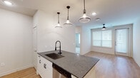 This single-story residence offers a thoughtfully designed open-concept layout connecting the kitchen and living areas. The kitchen is a standout feature, showcasing a central island with dark granite countertops, an undermount sink paired with a matte black spring-neck faucet, and a stainless steel dishwasher below. White shaker-style cabinetry with black hardware lines the walls, complemented by a subway tile backsplash and a full suite of stainless steel appliances including a refrigerator, range, and over-the-range microwave. Three industrial-style pendant lights hang above the island, enhancing the contemporary feel. The adjoining living area is finished with warm wood-look flooring, a ceiling fan, and generous windows fitted with horizontal blinds that invite abundant natural light. A dedicated bedroom features soft carpeting, a ceiling fan, and a large window with outdoor views. The bathroom is well-appointed with a granite-topped vanity, white cabinetry, and a large framed mirror with bar lighting above. A separate laundry room houses a full-size washer and dryer, adding practical convenience throughout this clean, modern space.