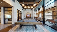 This expansive single-floor amenity space showcases a compelling blend of industrial and rustic design elements unified by whitewashed brick walls, dark hardwood flooring, and soaring double-height ceilings. The centerpiece of the space is a generously sized game room anchored by a live-edge wood billiard table set atop a vintage-style area rug, illuminated by a cluster of industrial pendant lights suspended from exposed wood beam coffers with a dark ceiling finish.

Adjacent to the game room, a sophisticated lounge area features floor-to-ceiling built-in shelving with warm accent lighting, upholstered seating, and window bench seating that overlooks an outdoor pool courtyard, flooding the space with natural light. A dedicated library nook provides a quiet retreat with lit shelving, leather chairs, and additional courtyard views.

A glass-walled conference room, enclosed by black steel-framed partitions with draping curtains, features a long communal table beneath Edison-style pendant lights and a wood plank ceiling. A private study offers a live-edge wood desk and backlit shelving against exposed brick. The kitchen is outfitted with Viking appliances, warm wood cabinetry, white subway tile, and farmhouse-inspired décor, completing the space with practical elegance.