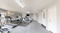 Located at 6024 N Jefferson St in Kansas City, Missouri, this fitness center offers a clean, functional space designed to support a well-rounded exercise routine. The room features durable speckled concrete flooring throughout, with rubber workout mats positioned beneath the equipment for added stability and safety. Bright fluorescent lighting is mounted to the textured ceiling, ensuring the space is well-illuminated at all times.

The exercise room is equipped with a variety of cardio and strength-training machines, including treadmills, an elliptical, a stationary bike, and an adjustable weight bench. A rack of dumbbells and additional free weights provide options for resistance training. A wall-mounted flat-screen television is positioned for easy viewing during workouts, accompanied by a standing fan for ventilation. A water cooler is conveniently available within the room.

A large wall mirror on one side of the room visually expands the space and allows users to monitor their form. On the opposite end, a set of French doors floods the room with natural light and offers views of the outdoor surroundings, creating an open and inviting atmosphere. A bulletin board and a small rack of exercise accessories, such as resistance bands and mats, round out the practical amenities. Multiple access doors provide convenient entry and exit points throughout the space.