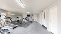 Located at 6024 N Jefferson St in Kansas City, Missouri, this fitness center offers a clean, functional space designed to support a well-rounded exercise routine. The room features durable speckled concrete flooring throughout, with rubber workout mats positioned beneath the equipment for added stability and safety. Bright fluorescent lighting is mounted to the textured ceiling, ensuring the space is well-illuminated at all times.

The exercise room is equipped with a variety of cardio and strength-training machines, including treadmills, an elliptical, a stationary bike, and an adjustable weight bench. A rack of dumbbells and additional free weights provide options for resistance training. A wall-mounted flat-screen television is positioned for easy viewing during workouts, accompanied by a standing fan for ventilation. A water cooler is conveniently available within the room.

A large wall mirror on one side of the room visually expands the space and allows users to monitor their form. On the opposite end, a set of French doors floods the room with natural light and offers views of the outdoor surroundings, creating an open and inviting atmosphere. A bulletin board and a small rack of exercise accessories, such as resistance bands and mats, round out the practical amenities. Multiple access doors provide convenient entry and exit points throughout the space.