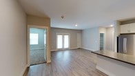 This single-floor residence offers a clean, modern layout designed for comfortable everyday living. The main living area is anchored by light-toned hardwood-style flooring that extends throughout the open space, creating a bright and cohesive aesthetic. Neutral gray and beige walls provide a versatile backdrop, complementing the contemporary finishes found throughout the home. A set of French doors along the back wall floods the living area with natural light while offering access to the outdoors, enhancing the sense of openness and airflow. Recessed lighting in the ceiling adds a polished touch and ensures well-distributed illumination throughout the day and evening. A glimpse into the adjacent room reveals plush carpeting and a window, suggesting a comfortable bedroom or private retreat. To one side, a built-in shelving area provides practical storage, while a partial view of the kitchen counter hints at a connected, functional kitchen space nearby. White trim and baseboards throughout lend a crisp, finished look to each room. The overall ambiance is calm and inviting, with a thoughtful, open floor plan that balances practicality with a welcoming residential atmosphere suited for a variety of lifestyles.