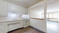 This single-floor unit offers a straightforward and functional layout designed for comfortable everyday living. The living area is bright and open, featuring wood-look hard flooring, neutral white walls, and a large window that allows natural light to fill the space. A wall-mounted HVAC unit provides climate control beneath the window. The kitchen is well-equipped with white cabinetry extending to the upper walls, a double sink, electric range with overhead ventilation, and a full-size refrigerator. A pass-through window with a wooden ledge connects the kitchen to the living area, creating an open, communicative feel between the two spaces. The flooring throughout the kitchen and living areas is durable wood-look material, lending a cohesive aesthetic. The bathroom is finished with classic white square wall tiles, a mosaic tile floor, a soaking bathtub with a shower, a wall-mounted sink, a mirrored medicine cabinet, and a standard toilet. The bedroom features neutral carpet and clean white walls, offering a quiet and comfortable retreat. A closet is accessible from the central hallway, which also connects the various rooms of the unit in an efficient and organized manner.