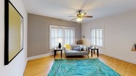 This single-floor unit showcases consistent hardwood flooring throughout, lending warmth and continuity across all rooms. The living room is a welcoming and well-lit space, featuring multiple white-framed windows that allow generous natural light to fill the room. A ceiling fan with a classic brass finish anchors the space from above, while a baseboard heating system ensures year-round comfort. A distinctive interior pass-through window with a wooden sill adds a charming architectural detail connecting the living area to adjacent spaces.

The kitchen features warm wood cabinetry with black hardware, a double stainless steel sink set in a light granite-style countertop, and a white refrigerator. A bright breakfast nook adjacent to the kitchen offers a cozy dining spot with its own window.

Both bedrooms are comfortably sized, each equipped with a ceiling fan and windows providing natural light. The rooms feature neutral wall tones and the same hardwood flooring found throughout the unit.

The bathroom is a standout, showcasing a classic clawfoot tub with an overhead shower curtain rod, original hex tile flooring, white wainscoting, a vintage cast-iron radiator, and a window that brings in natural light, creating a charming, character-filled atmosphere.