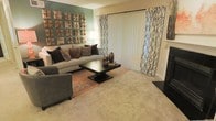 This thoughtfully designed single-floor space makes a strong first impression with its well-appointed living room. The room centers around a plush, neutral-toned sectional sofa adorned with an array of decorative throw pillows in rich earth tones, creating a warm and inviting atmosphere. A sleek, dark coffee table anchors the seating arrangement atop a patterned area rug that adds texture and visual interest to the soft carpeted flooring. One wall serves as a striking focal point, painted in a deep teal hue and adorned with a grid of sculptural metallic wall art, lending an artistic and contemporary flair to the space. Patterned drapery panels flank a set of vertical blinds covering a large sliding door, which allows for adjustable natural light and hints at outdoor access. A floor lamp provides warm ambient lighting, complementing the overall cozy yet modern aesthetic. The neutral carpeting flows throughout, contributing to a cohesive and comfortable feel. The space strikes a balance between bold design choices and functional living, making it both visually dynamic and livable.