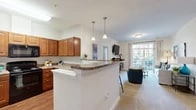 Located at 7500 Hearthside Way in Elkridge, MD, this well-appointed two-bedroom, two-bathroom residence offers a thoughtfully designed single-floor layout. The open-concept kitchen is outfitted with warm wood cabinetry, granite-style countertops, black appliances, and hardwood-style flooring. Pendant lights hang above a peninsula island that transitions seamlessly into the dining area and the carpeted living room beyond. The living room is bright and inviting, with large sliding glass doors that fill the space with natural light and open to a private balcony with outdoor views. A dedicated dining area features rich hardwood-style flooring and track lighting, providing a distinct yet connected space for entertaining. The primary bedroom is generously sized, with plush carpet, natural light from large windows with blinds and drapes, and ample closet space. The second bedroom offers a cozy, versatile layout ideal for guests or a home office. Both bathrooms are finished with wood-toned vanities and tub-shower combinations with clean white tile surrounds. Two closets provide practical storage throughout. The overall ambiance is bright, clean, and contemporary, with a neutral color palette that lends warmth and cohesion across the entire residence.