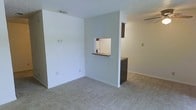 This single-floor residence offers a clean, functional layout designed for comfortable everyday living. The living area is finished with soft, neutral-toned carpeting that adds warmth underfoot, while the walls are painted in a light, airy palette that maximizes the sense of space and brightness throughout.

A standout architectural detail is the pass-through window cut into the dividing wall between the living area and the kitchen. This feature enhances connectivity between the two spaces, allowing for easy communication and flow while maintaining a defined separation. The kitchen area, visible through the pass-through, includes dark cabinetry that provides a subtle contrast to the lighter surroundings.

The layout is open and unfussy, with clean wall lines and simple baseboards that contribute to a tidy, minimalist aesthetic. Ceiling-mounted HVAC vents are integrated discreetly into the textured ceiling, ensuring climate comfort throughout. The space benefits from a neutral, move-in-ready condition that offers a versatile canvas for a variety of interior styles and furnishing arrangements. Overall, the residence presents a straightforward, well-maintained environment suitable for a range of occupants.