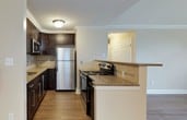 This single-floor residence offers a thoughtfully designed layout that balances comfort and functionality. The open-concept living area is anchored by warm hardwood-style flooring and flows seamlessly into the kitchen, which is outfitted with rich dark cabinetry, granite countertops, a tile backsplash, and a full suite of stainless steel appliances including a refrigerator, microwave, and range. A breakfast bar connects the kitchen to the living space, making it ideal for both everyday living and entertaining. Crown molding and recessed lighting add a refined touch throughout the common areas. The bedrooms feature plush carpeting and ample natural light; one standout bedroom showcases a dramatic vaulted ceiling paired with a striking arched window that fills the room with greenery views and abundant daylight. The two bathrooms are finished with light tile flooring, dark vanities topped with granite, and glass-enclosed showers — one featuring a tiled surround and the other a clean, minimalist design with a combination tub and shower. Neutral warm tones carried throughout the space create a cohesive and inviting ambiance from room to room.