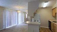 This single-floor space showcases an open-concept design that seamlessly connects the kitchen and living areas. The kitchen features a wraparound peninsula with a light countertop, white appliances including a coil-top range and refrigerator, and wood-toned upper cabinetry. A range hood is mounted above the stove, and the kitchen transitions smoothly into the living space via the peninsula's pass-through design. The living area is finished with wall-to-wall carpeting and is anchored by a decorative chandelier that adds a classic touch. A full-length mirrored wall enhances the sense of space and reflects natural light throughout the room. Large windows dressed with vertical blinds run along the exterior wall, allowing generous amounts of daylight to fill the interior. The ceilings feature a textured finish, and the transition between the kitchen's hard flooring and the carpeted living area provides a subtle distinction between the two functional zones. Overall, the space presents a practical and light-filled layout suited to everyday living.