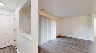 This single-floor space offers a thoughtfully arranged layout with a clean, modern aesthetic throughout. The living area is a standout feature, characterized by wood-look plank flooring in a warm gray tone and a large sliding glass door dressed with vertical blinds, allowing natural light to fill the room while providing direct views of the lush greenery outside. A pass-through cutout in the adjoining wall adds an open, connected feel between the living space and adjacent areas.

The galley-style kitchen is efficiently designed with two parallel runs of cabinetry finished in a light gray tone with sleek bar-pull hardware. White countertops provide a crisp contrast, complemented by a classic subway tile backsplash. Stainless steel appliances, including a refrigerator, electric range, and dishwasher, complete the functional setup. Matching wood-look flooring carries through from the living area into the kitchen, maintaining visual continuity.

The bedroom features neutral carpet flooring and a window fitted with horizontal blinds, offering a quiet, comfortable retreat with natural light. The bathroom presents a white shaker-style vanity with under-cabinet storage, a large mirror, and a well-lit vanity fixture above, all set against wood-look flooring for a cohesive finish throughout the space.