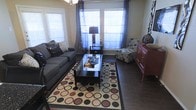 This single-story living space features a cozy and thoughtfully arranged sitting area centered around a bold geometric-patterned area rug in cream, black, and burgundy tones. Dark hardwood flooring extends throughout, providing a rich contrast to the lighter walls and furnishings. A gray upholstered sofa anchors one side of the room, complemented by a floral accent chair and a dark wood coffee table with a glass top. Natural light flows generously into the space through multiple windows dressed with white blinds and sheer blue curtains, creating a bright and welcoming atmosphere. A dark side table with a decorative lamp adds warmth and function between seating areas. On one side of the room, a red wooden cabinet provides both storage and a pop of color, accented by decorative pieces on top. The overall ambiance of the space is casual yet curated, with a mix of textures and tones that give the room a lived-in, comfortable character. The layout is efficient and open, making good use of the available area while maintaining clear pathways and a balanced visual flow throughout.