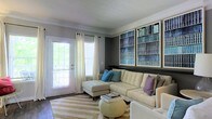 This two-story space includes a thoughtfully designed living area that combines comfort with visual interest. The room is anchored by a large, cream-colored sectional sofa adorned with teal, lavender, and neutral throw pillows, creating an inviting and relaxed atmosphere. A striped area rug in beige tones defines the seating area, complemented by a round, tufted ottoman that doubles as a coffee table.

One of the most striking features of the living area is the wall of framed built-in bookshelves, finished with crown molding and filled with neatly arranged books in cool blue and green tones, adding both a decorative and intellectual character to the space. The walls are painted in a cool gray tone, enhancing the room's modern yet cozy feel.

Natural light enters through a white-framed exterior door with glass panels and an adjacent window, both dressed with sheer white curtains that soften the light. Crown molding runs along the ceiling, adding an elegant architectural detail throughout. Dark hardwood flooring grounds the room, providing a warm contrast to the lighter furnishings and wall tones.