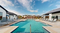 Located at 4005 N Elm St in Denton, Texas, this outdoor amenity complex centers around a generously sized resort-style swimming pool that serves as the focal point of the community. The pool area is designed with a modern aesthetic, featuring clean geometric lines, a wide surrounding deck finished in light-toned stone pavers, and an abundance of lounge chairs and chaise lounges arranged to maximize sun exposure throughout the day. Shallow entry steps and multiple pool access points make the space inviting and accessible. A separate spa or wading area is integrated into the pool layout, adding to the resort-like atmosphere. The clubhouse building adjacent to the pool is constructed in a contemporary farmhouse style, characterized by white horizontal siding, gray brick accents, a pitched dark-shingled roof, and large picture windows that allow views across the pool deck. The building provides shaded covered patio areas outfitted with seating and umbrellas, offering a comfortable retreat from the Texas sun. The surrounding residential community is visible in the background, providing a cohesive neighborhood setting. The overall ambiance is bright, open, and welcoming, with a clear blue sky and well-maintained landscaping enhancing the outdoor living experience.