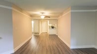 This single-floor space offers a clean, open layout that seamlessly connects the main living area to the kitchen. The flooring throughout is a warm-toned hardwood-style material that adds a sense of continuity and warmth across the space. Walls are finished in a soft neutral tone, complemented by crisp white baseboards and crown molding that frame the room with a polished, classic look.

A ceiling fan with a light fixture is centrally positioned, providing both air circulation and overhead illumination. The open sightlines from the living area extend toward a hallway that leads to additional rooms, lending a sense of depth despite the compact footprint.

The kitchen is visible from the living area and features white upper cabinetry, a light countertop, and under-cabinet space that allows for functional food preparation. A pass-through or peninsula-style counter partially separates the kitchen from the main living space while maintaining an open feel.

A mirrored element visible along one wall enhances the sense of space by reflecting light. A floor vent indicates a central HVAC system. Overall, the space presents a bright, tidy, and functional environment suited for everyday living.