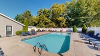 Located at 7670 N Oakbrook Dr in Reynoldsburg, Ohio, this two-floor community space centers around a generously sized outdoor swimming pool that serves as the focal point of the property. The pool area is enclosed by a white lattice privacy fence, creating a clean and welcoming atmosphere, while mature trees lining the perimeter provide natural shade and a lush green backdrop.

The pool deck is expansive, offering ample room for lounging with numerous blue reclining chaise lounge chairs and side tables arranged around the perimeter of the pool on all sides. The layout comfortably accommodates a large number of guests simultaneously. Entry into the pool is facilitated by built-in stainless steel handrails on multiple sides.

Adjacent to the pool is a brick and siding clubhouse building with a pitched shingle roof. The structure features large windows and glass-panel entry doors that open directly onto the pool deck, providing convenient indoor-outdoor access. The exterior brick facade gives the building a classic, well-maintained appearance, complemented by neatly trimmed shrubbery at its base.

Under clear blue skies, the entire outdoor space is flooded with natural sunlight, making it an inviting recreational area. The surrounding neighborhood views, including a nearby church steeple visible beyond the fence line, add to the community character of this well-appointed outdoor amenity space.