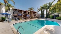 The Missions At Back Bay pool amenity area in Costa Mesa, California offers residents a well-appointed outdoor retreat designed for relaxation and recreation. The centerpiece of this space is a large, rectangular swimming pool with clear blue water, accessible via built-in steps and a handrail entry point. The expansive concrete pool deck is lined with blue sling-style chaise lounges and multiple large blue market umbrellas, providing ample space for sunbathing and shade. Surrounding the pool, the landscaping features mature palm trees, Italian cypress trees, and trimmed hedgerows that create a lush, private atmosphere. Seating areas with tables and chairs are tucked along the perimeter, offering spots for casual outdoor dining or socializing. The pool area is enclosed by wrought iron fencing, adding both security and an elegant aesthetic. The Spanish Mission-style residential buildings frame the courtyard on multiple sides, featuring stucco exteriors, red tile rooflines, and wood-railed balconies that overlook the pool. The overall ambiance is that of a sun-drenched Southern California retreat, blending functional amenities with a warm, resort-inspired architectural character under the consistently clear skies of the Orange County climate.
