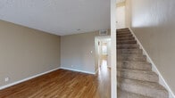 This two-story home in Pleasanton, CA (94566) offers a practical and comfortable layout spread across two levels. The main floor features warm wood-style flooring throughout the open living areas, which are bathed in neutral beige tones and finished with textured ceilings. A carpeted staircase with a railed banister connects the levels, with a utility closet neatly tucked beneath. The kitchen is well-appointed with rich wood cabinetry, dark granite-style countertops, stainless steel appliances including a refrigerator, range, and dishwasher, along with wood-style flooring that ties into the main living space.

The upper level transitions to plush carpeting, providing a softer feel in the bedroom areas. One bedroom features a large picture window with vertical blinds, allowing natural light and outdoor views, while another includes direct access to a generously sized walk-in closet with dual hanging rods and shelving on multiple walls. A bathroom with neutral walls and clean finishes completes the upper level. Central HVAC with ceiling vents serves both floors, ensuring year-round comfort throughout the home.