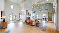 This single-floor space offers a generous and well-appointed layout spanning a variety of interconnected living areas. The entrance greets visitors with a striking herringbone brick floor, a sculptural round entry table, and soaring vaulted ceilings accented by sage-green exposed beams and a statement brass chandelier with woven pendant shades, establishing an immediate sense of architectural grandeur.

The great room is the heart of the space, featuring dramatic cathedral ceilings, light hardwood flooring, and a full-height shiplap accent wall anchored by a classic brick fireplace. Tall French doors flood the room with natural light and provide views of the outdoors, while clerestory windows further enhance the bright, airy atmosphere. Multiple seating arrangements and curated artwork create a refined yet inviting ambiance.

The kitchen is well-equipped with white shaker cabinetry, brushed gold hardware, quartz countertops, a subway-tile backsplash, stainless steel appliances, and light hardwood floors. A pass-through connects it seamlessly to the dining room, which features soft blue walls, a breakfast bar, and warm wood furnishings.

A separate living area, painted in a rich teal, offers a more intimate retreat with large windows, a coffered ceiling, a woven floral light fixture, and abundant natural light throughout.