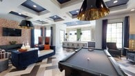 This sophisticated entertainment lounge is designed to impress, combining upscale aesthetics with functional recreational amenities. At the heart of the space sits a full-size billiards table, illuminated by a bold industrial-style pendant light with a gold interior that adds a glamorous touch overhead. The coffered ceiling, finished in deep navy blue with recessed lighting, creates a dramatic architectural focal point that draws the eye upward and adds a sense of grandeur to the room. Underfoot, a geometric herringbone-patterned floor in muted tones of gray and white adds visual depth and contemporary flair throughout the open layout. A stylish bar area anchors the rear of the space, complete with sleek bar stools, pendant lighting, and a shimmering mosaic tile accent wall that reflects light beautifully. Adjacent to the bar, a lush vertical green wall adds a refreshing natural element. The lounge seating area features plush high-back navy blue chairs accented with vibrant orange pillows, set against exposed brick walls and large windows that flood the space with natural light. Elegant navy drapes frame the windows, completing the polished, cohesive design of this well-appointed social space.