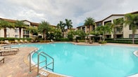 This inviting residential community showcases a beautifully designed outdoor amenity area centered around a generously sized freeform swimming pool with crystal-clear blue water. The pool deck is finished with tasteful pavers and equipped with stainless steel entry railings, providing safe and easy access. Surrounding the pool, a well-appointed lounging area features multiple patio umbrella-shaded seating arrangements, ideal for relaxing in the warm Florida-style climate. Mature, towering palm trees and lush tropical greenery frame the entire setting, creating a sense of seclusion and resort-like ambiance. The backdrop consists of multi-story residential buildings with warm earth-toned stucco exteriors, terracotta-style rooflines, and private balconies that overlook the pool courtyard. The architecture reflects a Mediterranean-inspired design aesthetic, with structured facades complemented by the natural softness of the surrounding landscape. The overall atmosphere of the outdoor space is serene and luxurious, evoking the feel of an upscale tropical retreat. The combination of well-maintained landscaping, generous poolside amenities, and thoughtfully designed architecture creates a cohesive and welcoming community environment suited for leisure and relaxation.