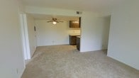 This single-floor residential space offers a practical and functional layout designed for comfortable everyday living. The main living area is generously sized, featuring wall-to-wall carpet flooring in a neutral beige tone that adds warmth throughout. A ceiling fan with an integrated light fixture is centrally mounted, providing both air circulation and ambient lighting to the open space. The living area flows seamlessly into the kitchen, which is partially separated by a half-wall partition, creating an open yet defined layout. The kitchen features dark wood cabinetry on both the upper and lower sections, offering ample storage, along with light-colored countertops that provide a clean contrast. A built-in appliance, likely a dishwasher, is visible beneath the counter. The walls throughout are painted in a clean, neutral white, contributing to a bright and airy atmosphere. Ventilation is supported by ceiling-mounted HVAC registers. The overall design is straightforward and utilitarian, making the space adaptable for a variety of furnishing styles. The open floor plan between the living and kitchen areas promotes a sense of spaciousness and ease of movement throughout the home.