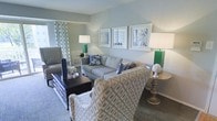 This single-floor space offers a cohesive and inviting interior that blends comfort with refined style. The living area serves as the centerpiece of the space, showcasing a carefully curated coastal-inspired design aesthetic. Soft, muted blue-gray walls create a calm and airy backdrop, complemented by a plush gray sofa accented with navy and floral throw pillows. Two patterned accent chairs — one featuring a classic geometric diamond print and another in a subtle gray weave — frame the seating arrangement, adding visual depth and texture. A dark espresso coffee table anchors the grouping, providing both function and contrast. A trio of framed art prints in coordinating blue tones lines the wall above the sofa, reinforcing the space's cohesive color story. Vibrant emerald green table lamps on either side of the room introduce a bold, energizing accent while maintaining the overall serene atmosphere. Patterned curtains with circular motifs dress the windows, allowing natural light to filter softly into the room. The flooring features a textured area rug that ties the furnishings together seamlessly. Overall, the space exudes a polished, resort-inspired ambiance that feels both welcoming and elegantly appointed.