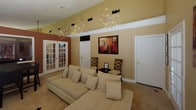 This single-floor space is defined by its impressive vaulted ceilings, which lend a sense of openness and grandeur to the interior. The living area serves as the focal point, featuring warm beige and terracotta-toned walls that create an inviting, comfortable atmosphere. A large sectional sofa in a neutral cream upholstery anchors the room, complemented by accent seating and small side tables.

A striking starburst chandelier hangs overhead, serving as a dramatic decorative centerpiece that enhances the height of the vaulted ceiling. Multiple large windows fitted with wood-tone horizontal blinds allow generous amounts of natural light to filter in while maintaining privacy. The flooring throughout transitions between light hardwood and classic travertine tile, adding visual texture and continuity.

A flat-screen television is mounted on a media console, while French doors and interior doorways connect the space to adjoining areas. A compact dining set with dark wood chairs occupies a defined dining zone, partially separated by an architectural pass-through. Bold abstract artwork on the walls adds color and personality. Overall, the space balances casual comfort with refined design details.