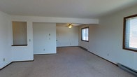 This single-floor residence offers a straightforward and functional layout designed for comfortable everyday living. The main living area is generously sized, featuring neutral wall-to-wall carpeting that creates a warm and cohesive feel throughout the space. White walls provide a clean, bright backdrop that lends itself well to a variety of décor styles. A ceiling fan is centrally positioned overhead, offering both lighting and air circulation. A window along one wall allows natural light to filter into the room, contributing to an airy atmosphere. Baseboard heating runs along the perimeter of the living area, ensuring efficient warmth throughout the colder months. Subtle architectural details, including a low-profile beam that partially divides the open area, add a hint of structure without disrupting the overall flow of the space. Light switch panels and electrical outlets are conveniently placed throughout, reflecting a practical and user-friendly design. The open layout offers flexibility in how the space can be arranged and utilized, making it adaptable to a range of lifestyle needs. Overall, the space presents a clean, move-in-ready environment with a simple and functional aesthetic.